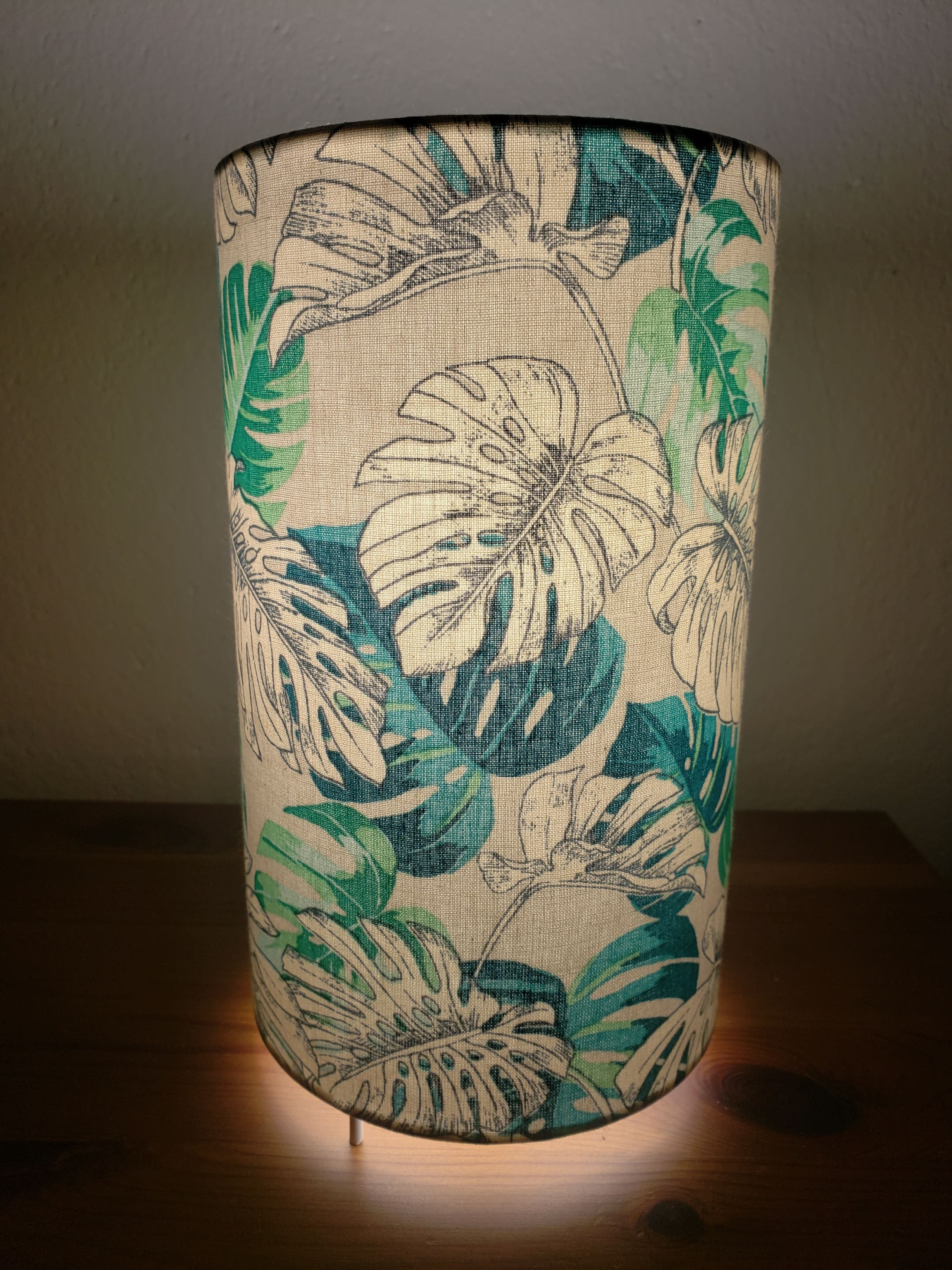 Photo d'une lampe en tissu cylindrique gris clair avec en motif de larges feuilles vertes et blanches 